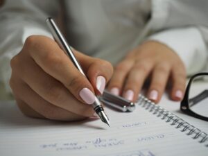 Woman-in-White-Long-Sleeved-Shirt-Holding-a-Pen-Writing-on-a-Paper